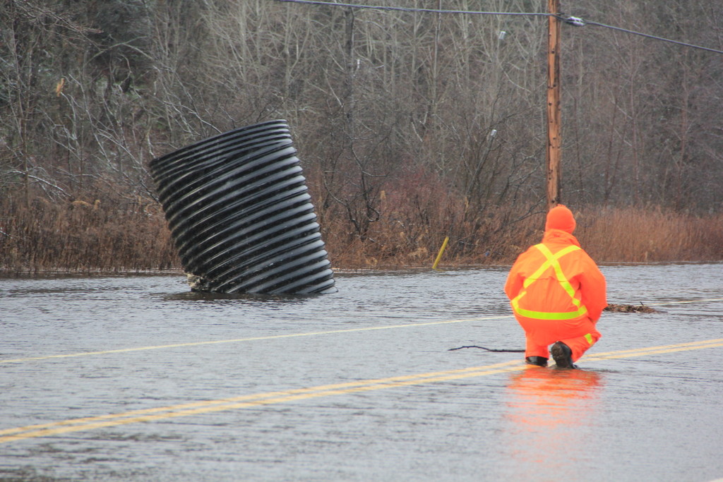 Worker in orange rain gear crouched on a flooded road beside a dislodged culvert
